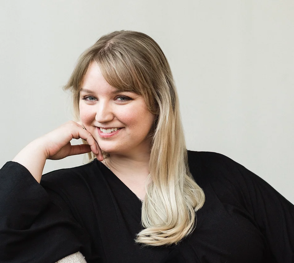 Mahala Bloom, owner of Bloom Studio, smiles at the reader. She has blonde hair and is wearing a black dress. Her chin rests on her hand.